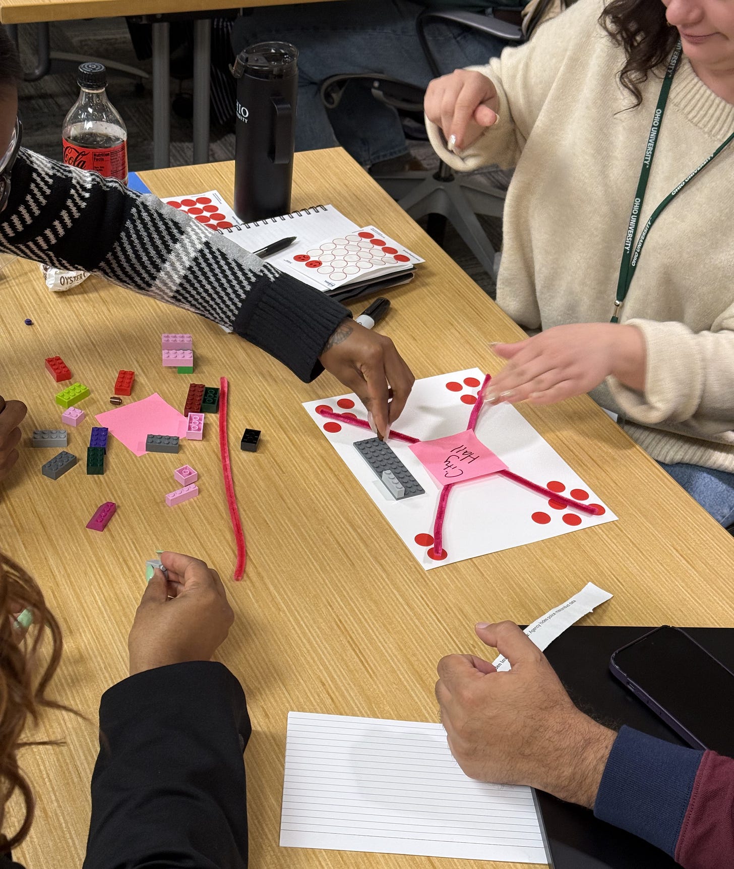 Kiplinger Fellows collaborate around a table during the Last Mile workshop, building a prototype utility tool using LEGOs, pipe cleaners, Post-it notes, and dot stickers. A hub-and-spoke model with a Post-it labeled 'City Hall' sits at the center of the workspace, with multiple hands reaching in to shape the design.