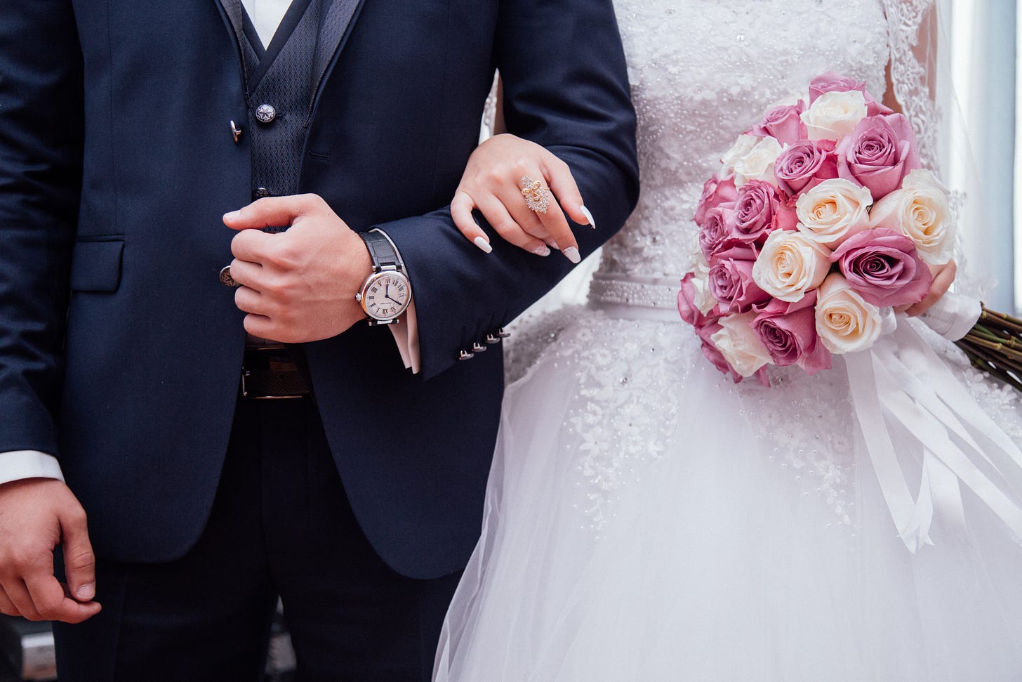 A bride and groom, hand-in-hand. The groom is wearing a dark suit and the bride is wearing a traditional, white wedding dress. She is holding a boquet of pink and white flowers.