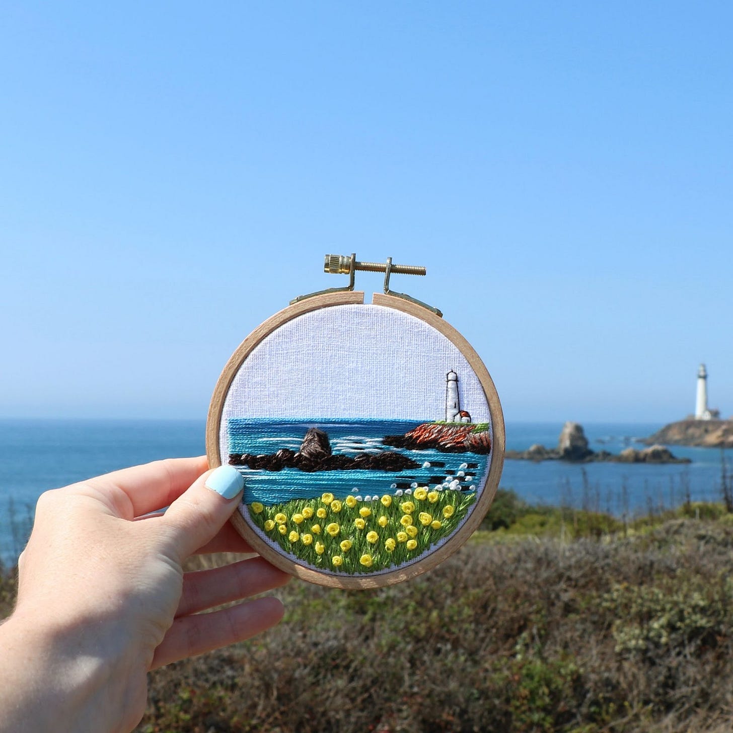 embroidered lighthouse hoop held up against coastal landscape