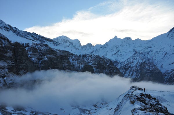 Long view of Annapurna mountain range in the cool dawn light. A person stands above the clouds looking at the abyss.  