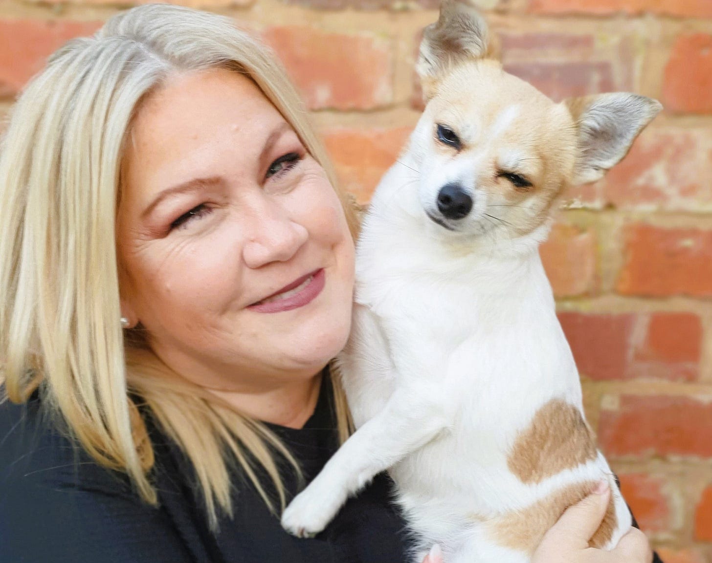 Smiling woman with straight blonde hair holds a small white and tan Chihuahua in front of a brick wall.