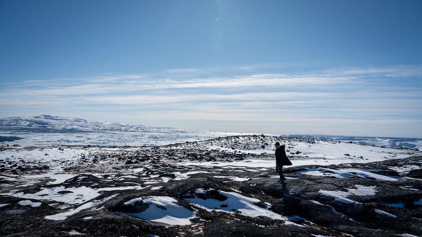 A lone figure standing on black rock and snow, looking out over a bay filled with icebergs near Ilulissat, Greenland. A lone figure standing on black rock and snow, looking out over a bay filled with icebergs near Ilulissat, Greenland.