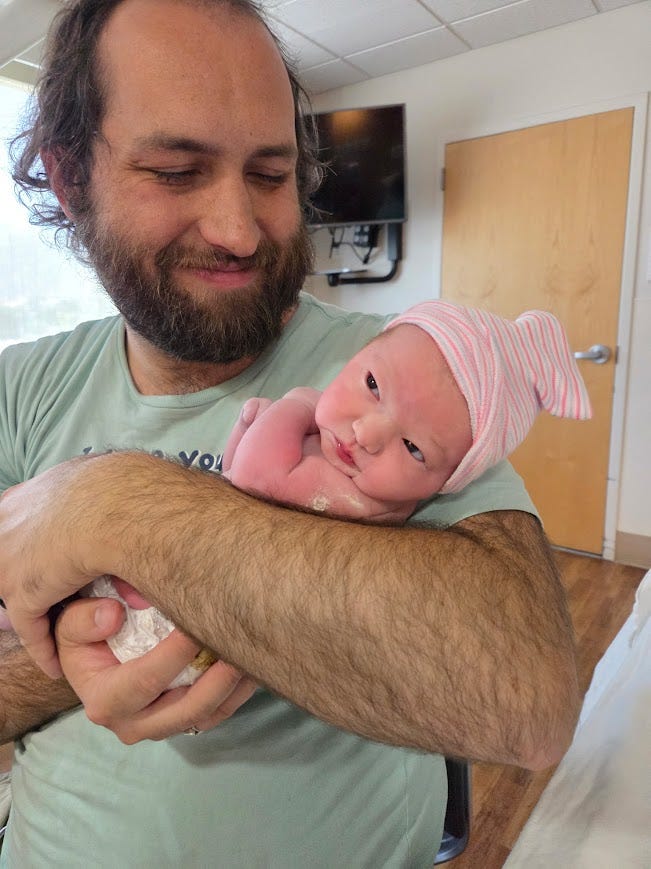 I'm standing holding a newborn baby, smiling down at her. She has a decidedly unenthused expression.