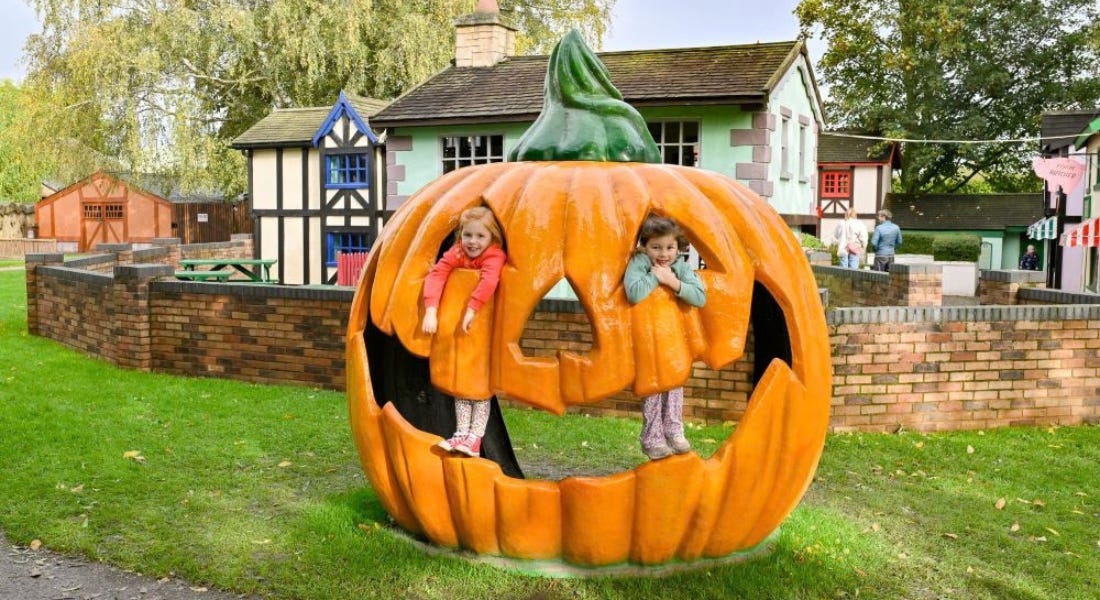 Two girls standing inside a pumpkin statue, with a group of houses behind them