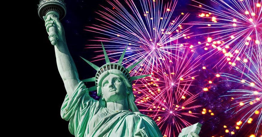Green bronze Statue of Liberty torso, one arm raised with torch, the other holding a book. in the background fireworks