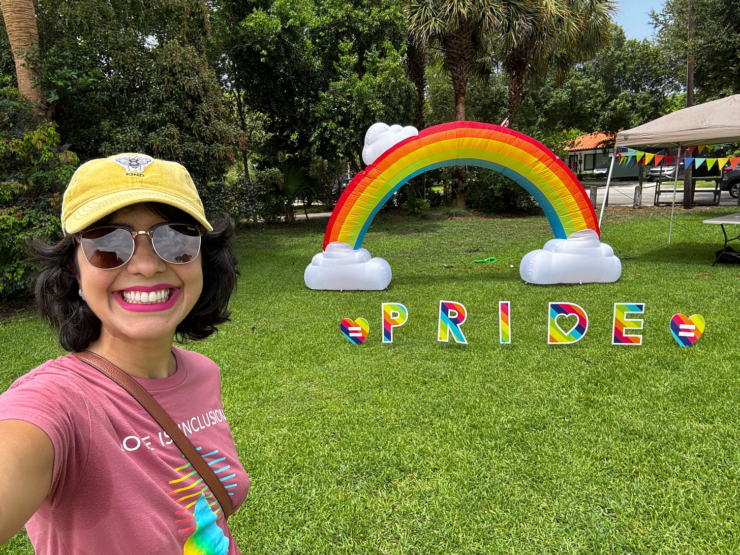 ALT Text: A smiling woman with dark hair wearing sunglasses, a yellow baseball cap, and a pink "Love is Inclusive" t-shirt takes a selfie at an outdoor Pride event. Behind her is a large inflatable rainbow arch with white clouds at each end, and colorful "PRIDE" letters spelling out the word on a green grass lawn. Tents and trees are visible in the background of what appears to be a community park setting. Edit ALT Text em Português: Uma mulher sorridente de cabelos escuros usando óculos de sol, boné amarelo e camiseta rosa escrita "Love is Inclusive" tira uma selfie em um evento Pride ao ar livre. Atrás dela há um grande arco inflável do arco-íris com nuvens brancas em cada extremidade, e letras coloridas formando a palavra "PRIDE" sobre a grama verde. Tendas e árvores são visíveis ao fundo do que parece ser um parque comunitário. ALT Text: A smiling woman with dark hair wearing sunglasses, a yellow baseball cap, and a pink "Love is Inclusive" t-shirt takes a selfie at an outdoor Pride event. Behind her is a large inflatable rainbow arch with white clouds at each end, and colorful "PRIDE" letters spelling out the word on a green grass lawn. Tents and trees are visible in the background of what appears to be a community park setting. Edit ALT Text em Português: Uma mulher sorridente de cabelos escuros usando óculos de sol, boné amarelo e camiseta rosa escrita "Love is Inclusive" tira uma selfie em um evento Pride ao ar livre. Atrás dela há um grande arco inflável do arco-íris com nuvens brancas em cada extremidade, e letras coloridas formando a palavra "PRIDE" sobre a grama verde. Tendas e árvores são visíveis ao fundo do que parece ser um parque comunitário.