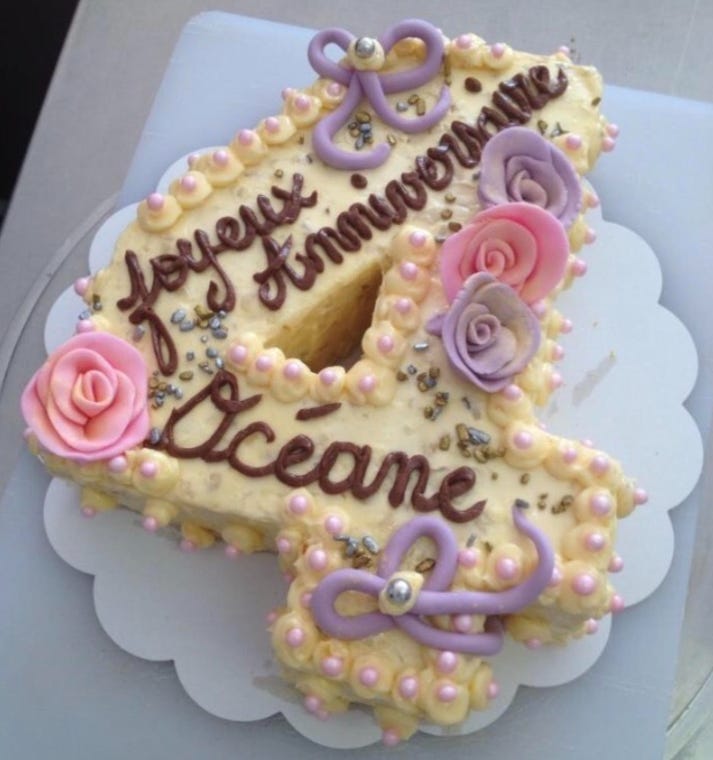 A birthday cake in the shape of the number four, covered with pale yellow icing and decorated with pink and purple roses and pale purple ribbons, all made of icing. The cake has brown, chocolate iced writing which reads Joyeux Anniversaire Océane A birthday cake in the shape of the number four, covered with pale yellow icing and decorated with pink and purple roses and pale purple ribbons, all made of icing. The cake has brown, chocolate iced writing which reads Joyeux Anniversaire Océane
