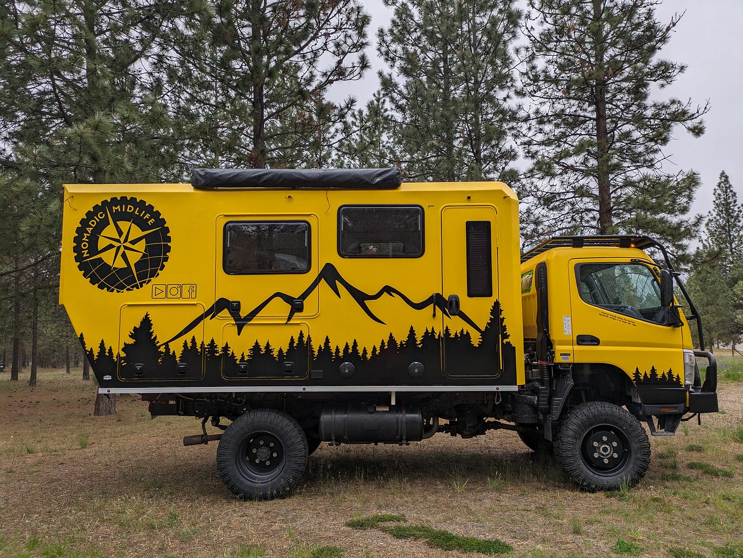 a big yellow camper truck built on a medium duty commercial chassis, decorated with black outlines of trees, mountain peaks, and a big logo that says Nomadic Midlife a big yellow camper truck built on a medium duty commercial chassis, decorated with black outlines of trees, mountain peaks, and a big logo that says Nomadic Midlife