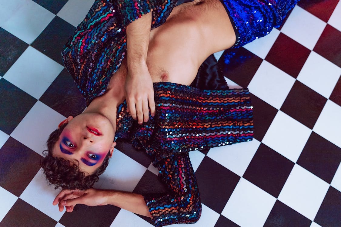 nonbinary young shirtless person in sparkly jacket and lots of makeup lying on checkerboard floor nonbinary young shirtless person in sparkly jacket and lots of makeup lying on checkerboard floor