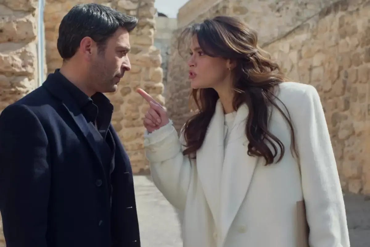 Uzak Şehir dizisinden bir sahne: Sinem Ünsal (Alya karakteri) beyaz bir palto giymiş, karşısındaki erkek oyuncuya (muhtemelen Cihan karakteri) parmağıyla işaret ederek güçlü bir tartışma anında görünüyor. Tarihi taş duvarlı bir mekânda çekilmiş.