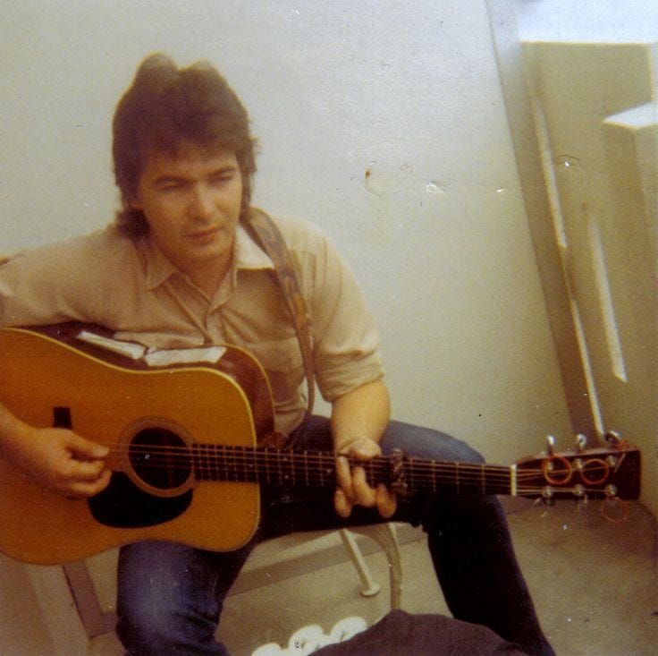 John Prine in his youth, holding a guitar and sitting on a chair. Folk music legend. Classic country legend.