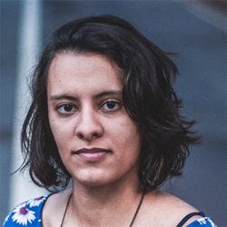 20251029_mila-pamplona-barbosa Uma mulher de cabelos na altura dos ombros, lisos e castanhos olha para a lente. Ela veste blusa azul com flores brancas e usa um colar longo.