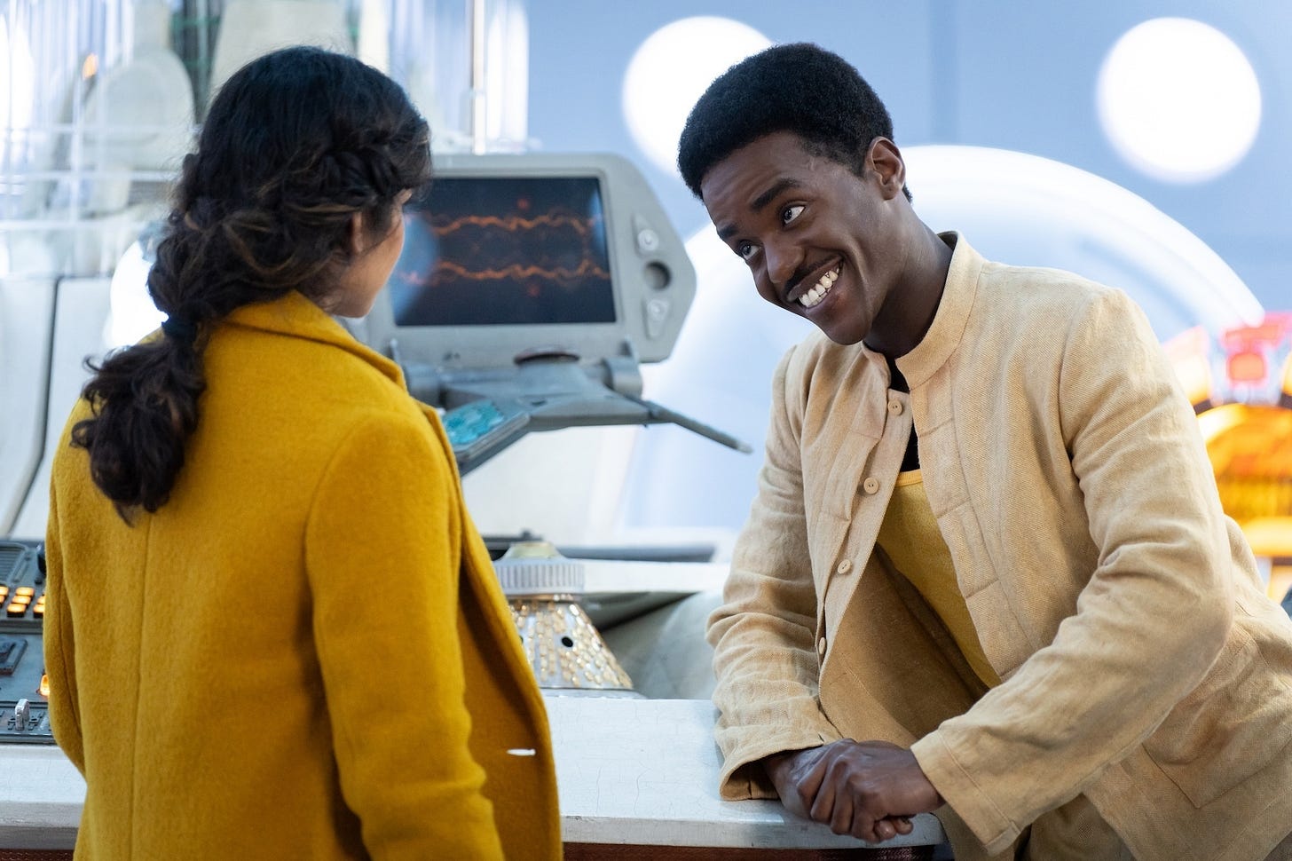 Cena do primeiro episódio da nova temporada de Doctor Who (segunda temporada) a personagem Belinda interperetada por Varada Sethu está de costas, olhando para o personagem Doctor interpretado por Ncuti Gatwa, homem negro, alto, mostrando um largo sorriso. Ao fundo telas e painéis de controle da nave espacial TARDIS