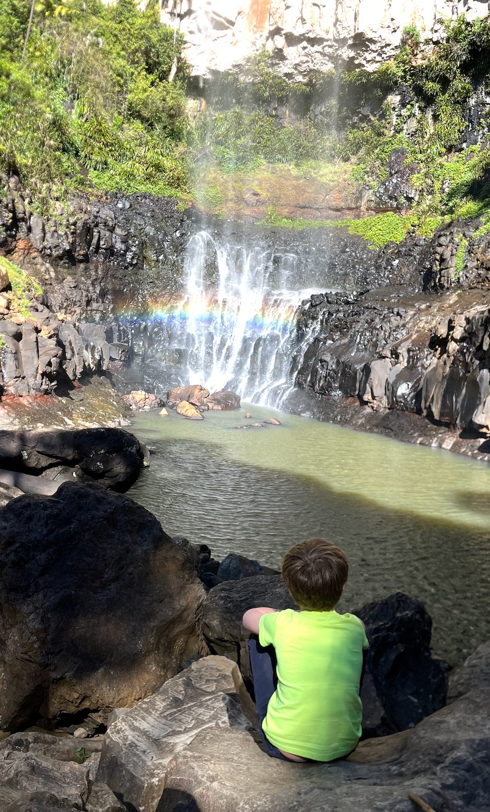 A boy sitting with his back to the camera looking at a waterfall that is creating a rainbow in it's mist.