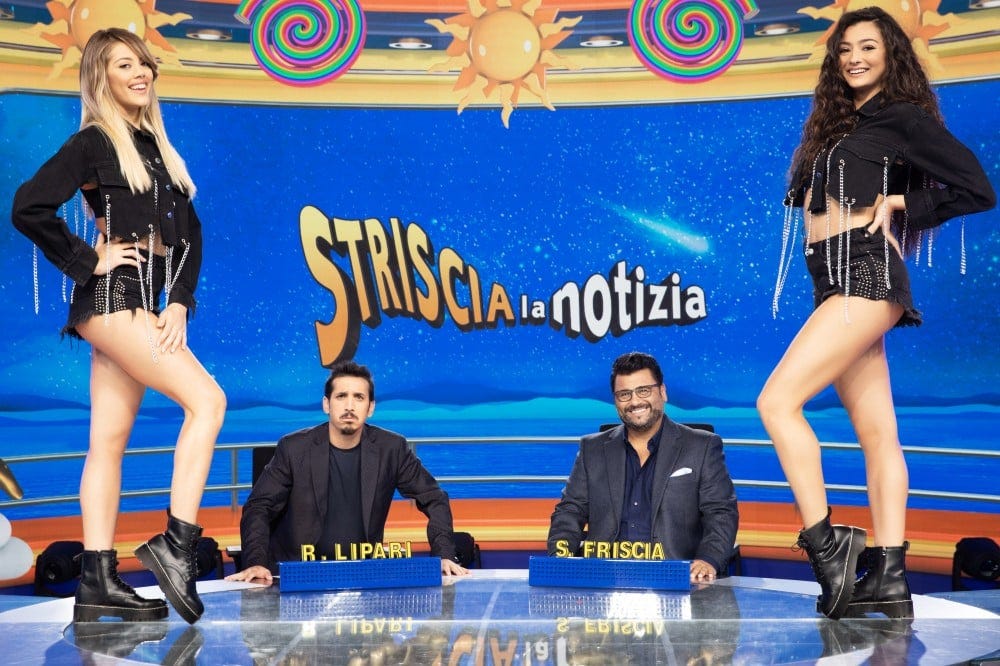 Immagine di Striscia la Notizia: conduttori seduti e veline sul tavolo, che mostrano gambe