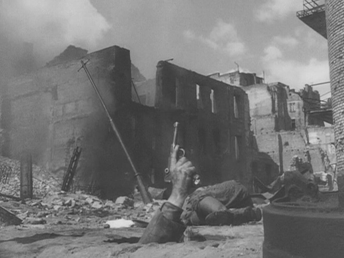 A hand clutching a pistol emerges from the ground, with the ruins of Warsaw in the background.