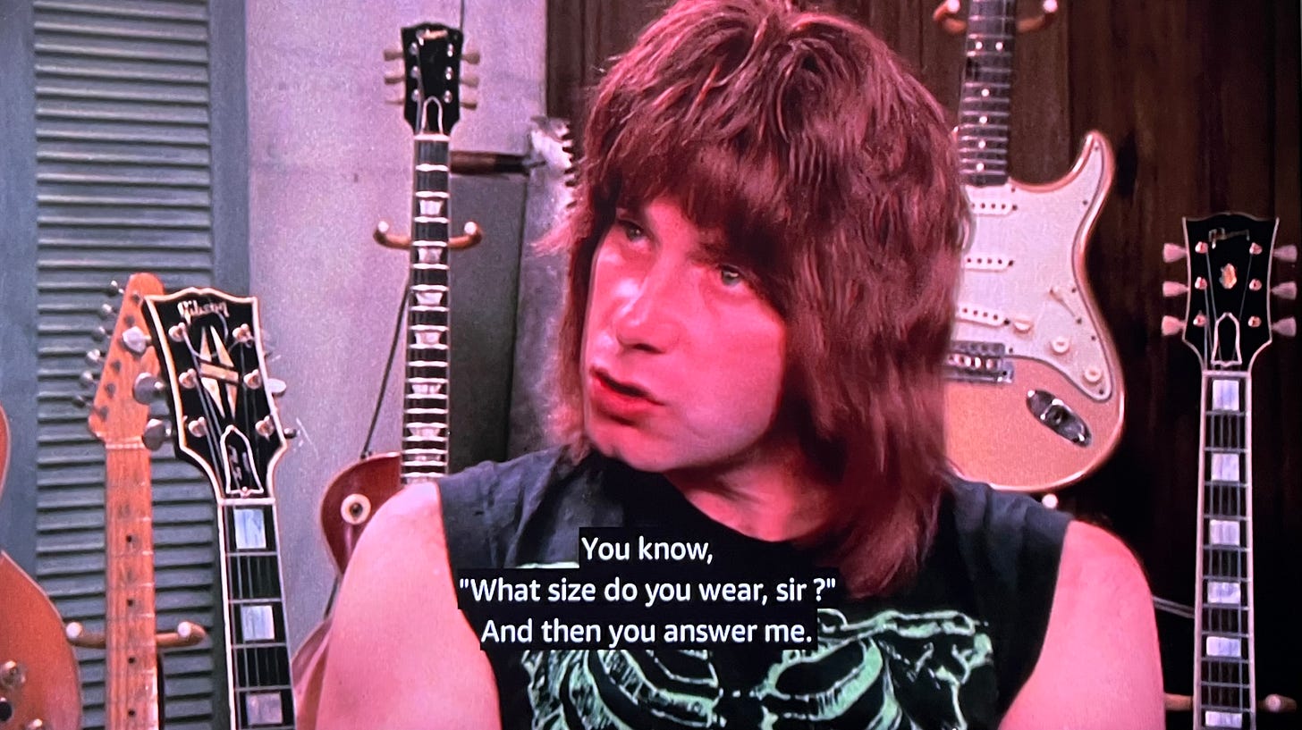Image from This is Spinal Tap: Nigel being interviewed by Marty diBergi in front of his guitars, and saying he could sell hats if he didn’t make music, like, “You know, ‘What size do you wear, sir?’ And then you answer me.” 