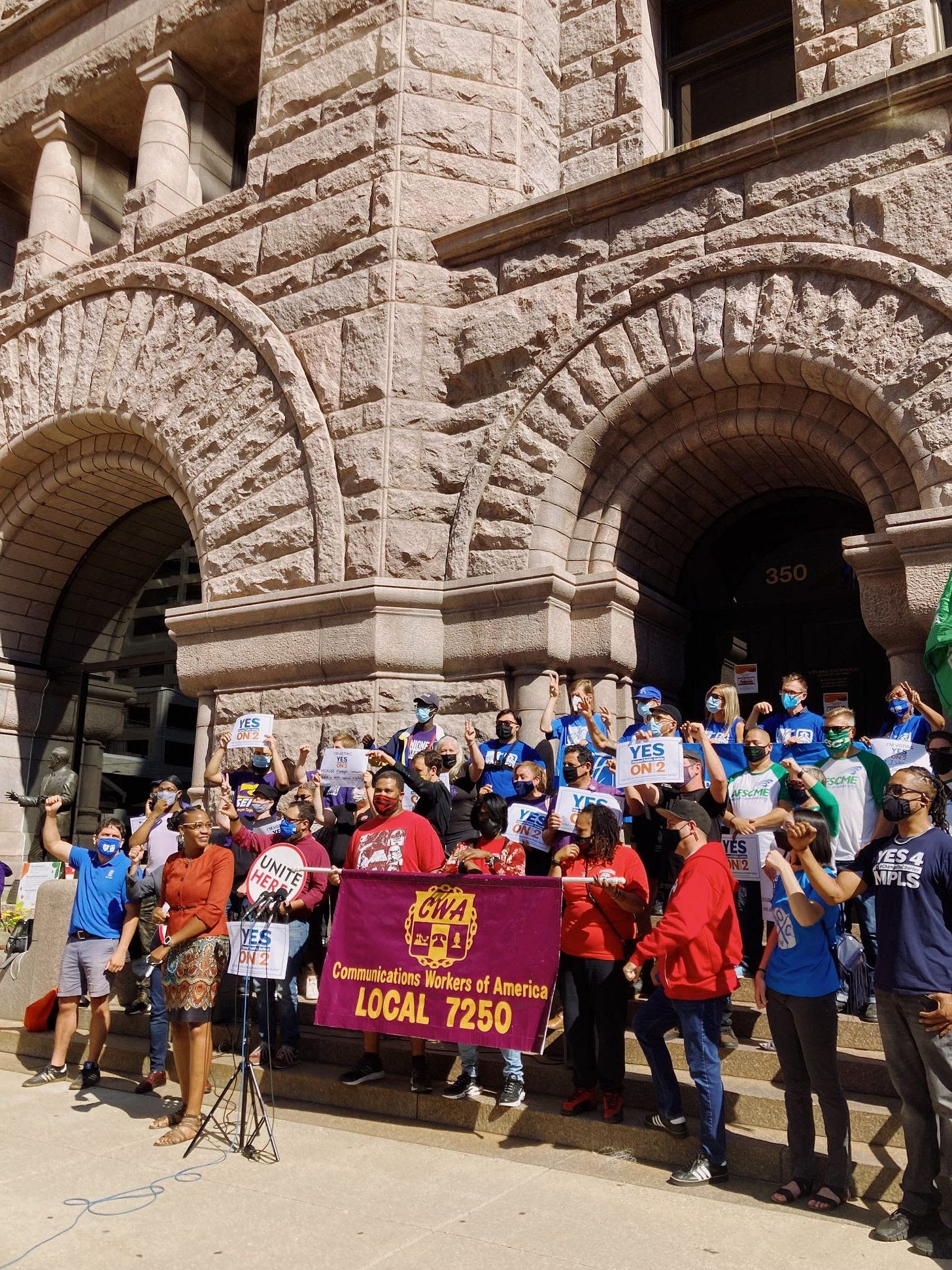 a group of people wearing masks and holding signs and fists up in front of beige/red city hall building steps