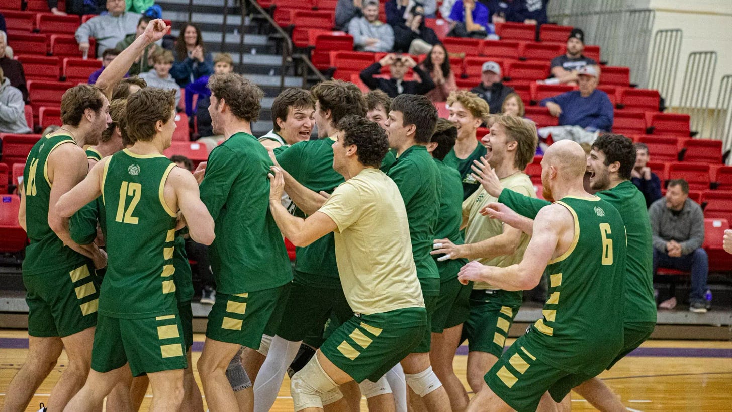 Members of the Missouri S&T men’s volleyball team celebrate together on the court following a point or match conclusion. The players, wearing green jerseys with gold accents and matching shorts, gather in a tight group, cheering, smiling, and raising their arms in excitement. Some players face inward toward teammates while others turn outward, capturing the energy of the moment. The scene takes place inside an indoor gymnasium with red bleacher seating in the background, where spectators are visible watching and reacting to the celebration.