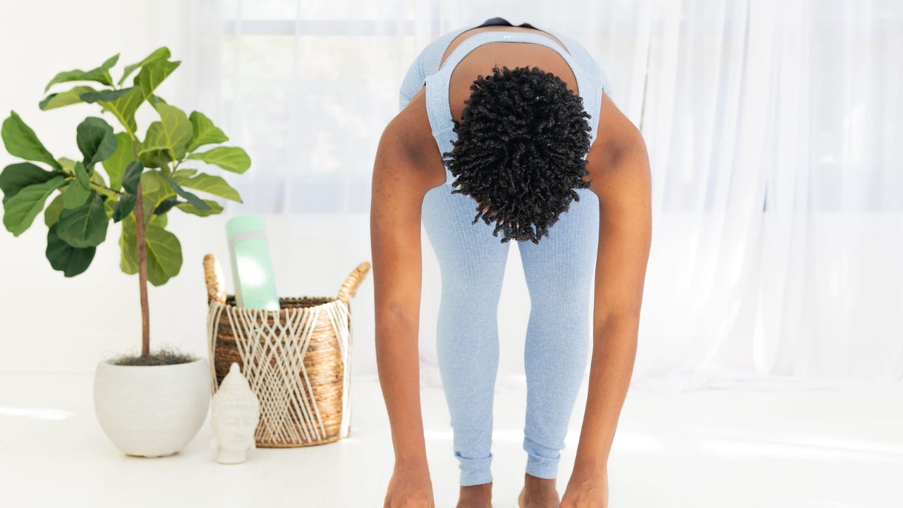 Black woman in light blue workout clothes bending forward in a standing stretch, with a yoga mat, plant, and basket visible in a bright home setting.
