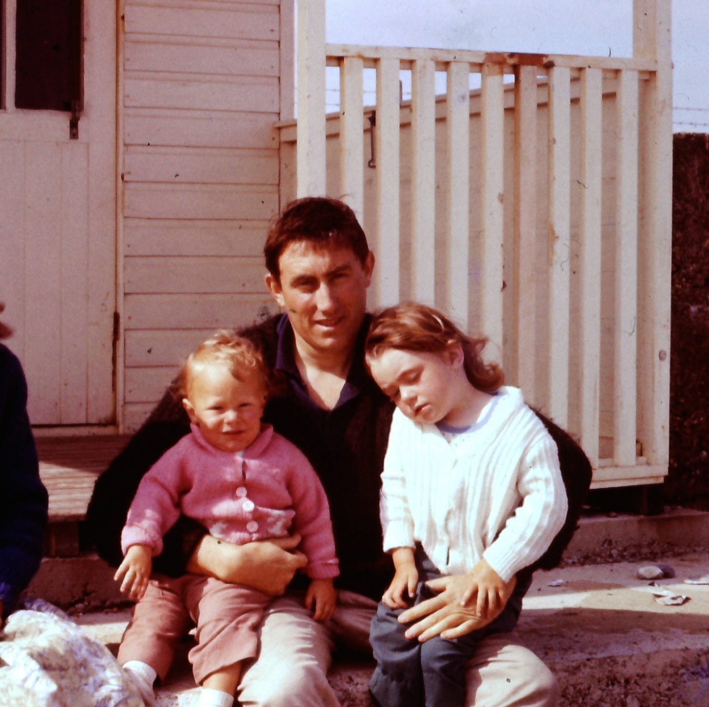 The photo shows Penny aged around four years old sitting on her dad Michael's lap while on the other side is her baby brother. Penny looks tired and keeps her hand firmly over her dad's. She wears a cute white cardigan and has blonde red hair. Her brother wears a pale pink cardigan with white buttons and little brown trousers. Michael is wearing a black jumper. He has black hair and a handsome face. They are sitting on the steps of a holiday chalet and the sun is out.