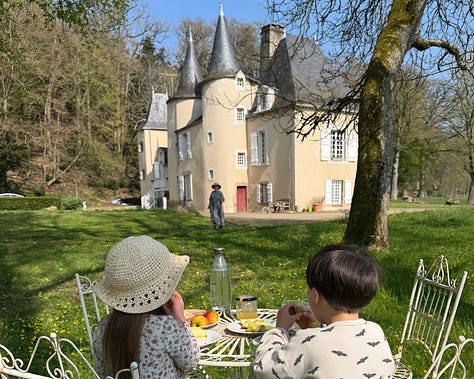 An overhead garden table with bread, cheese, carrots, apples, grapes and drinks; two children eating at a white bistro table with the château behind; two children running down a path toward the château.