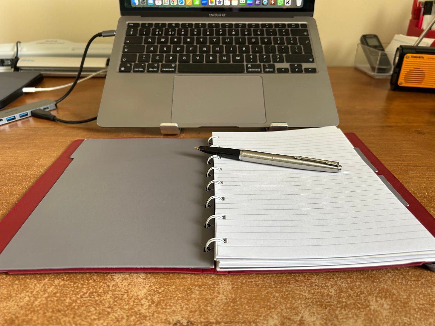 A notebook lies open on a desk. A fountain pen rests on the open notebook. Behind it is an Apple laptop, a Macbook Air. The notebook is a Filofax refillable in red. The pen a Parker, made in steel with a plastic end for the nib. Image by Roland Millward, Trowbridge, Wiltshire UK
