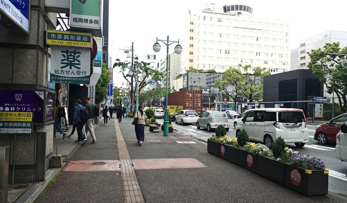 Street-level view of a Hakata (Fukuoka) sidewalk with yellow tactile paving down the centre, pedestrians walking past shopfronts with Japanese signage including a pharmacy, cars and a delivery truck on the road, and a flower planter box along the curb, with high-rise buildings in the background under an overcast sky. Street-level view of a Hakata (Fukuoka) sidewalk with yellow tactile paving down the centre, pedestrians walking past shopfronts with Japanese signage including a pharmacy, cars and a delivery truck on the road, and a flower planter box along the curb, with high-rise buildings in the background under an overcast sky.