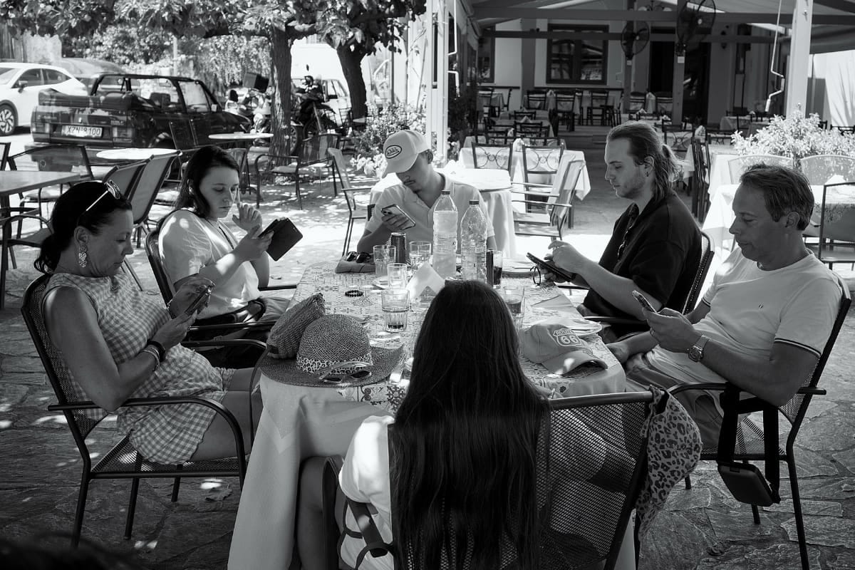 Black-and-white photograph of six people seated around a restaurant table in the shade, each absorbed in a smartphone, with drinks, hats and water bottles spread across the table.