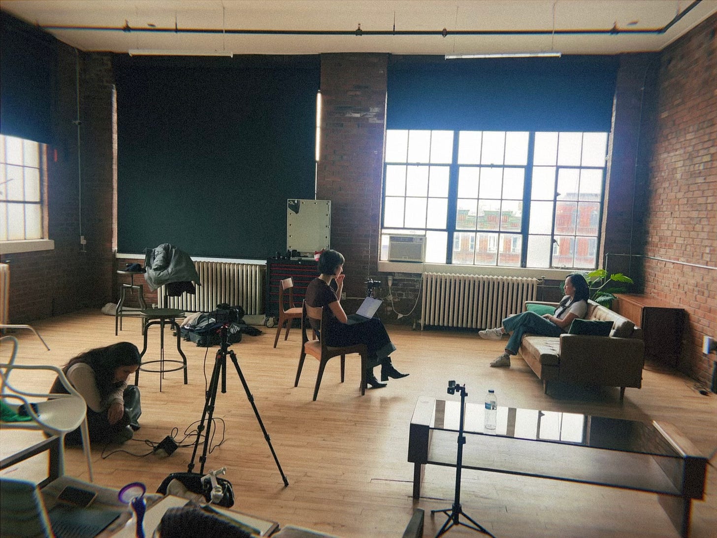 A studio setup with seating for conversation, natural light from large windows, and filming equipment arranged in an industrial loft space. A studio setup with seating for conversation, natural light from large windows, and filming equipment arranged in an industrial loft space.