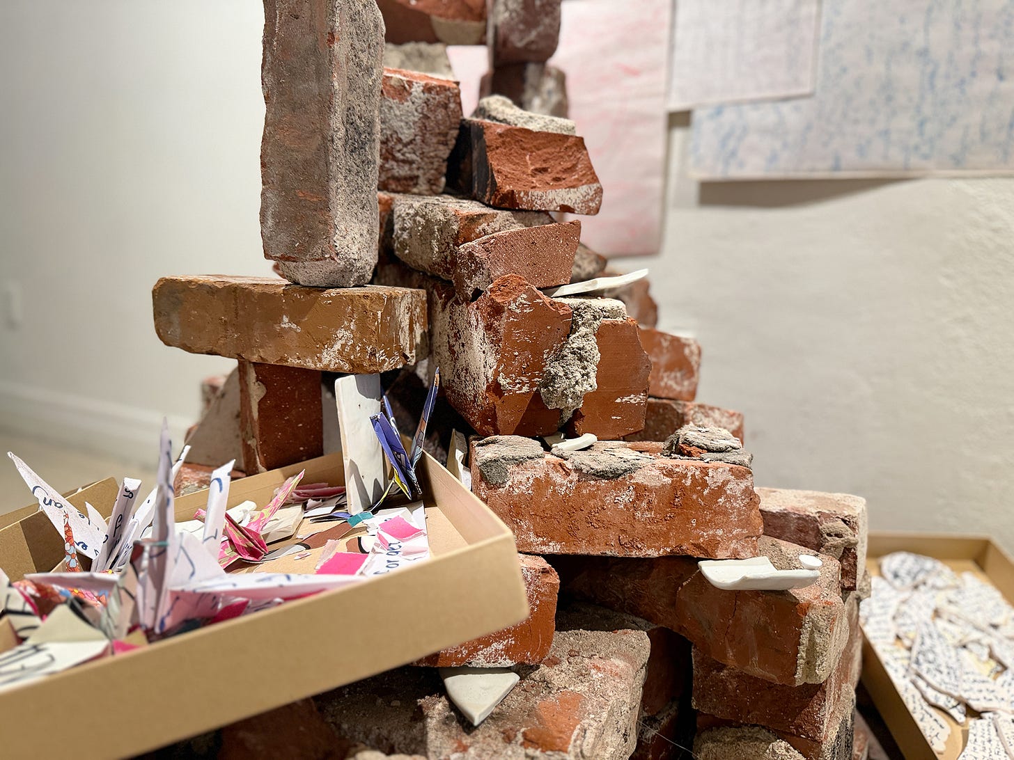 An open kraft-paper archive box sits among a pile of reclaimed bricks. Inside, folded paper cranes and small printed fragments. Handwritten notes are visible on the box lid.