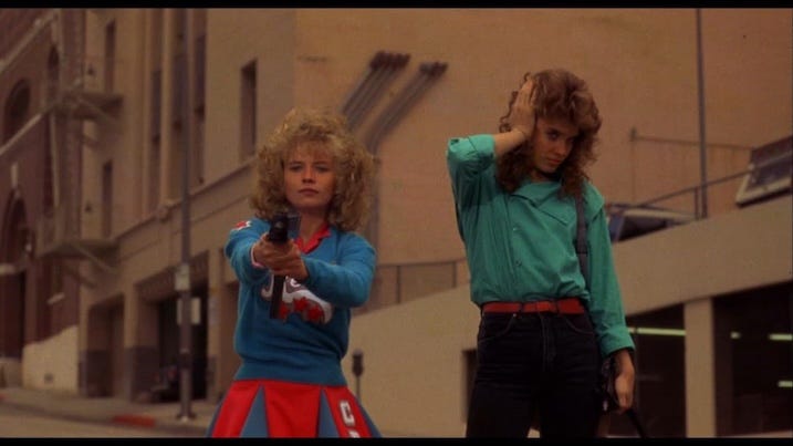 Still from Night of the Comet (1984) showing Kelli Maroney as Samantha aiming a gun while Catherine Mary Stewart as Regina stands beside her on an empty L.A. street.