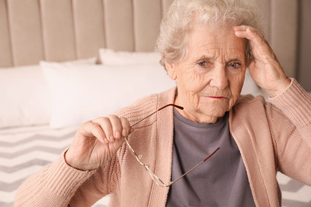 Senior woman in her bedroom, holding her glasses with one hand and her head with the other Senior woman in her bedroom, holding her glasses with one hand and her head with the other