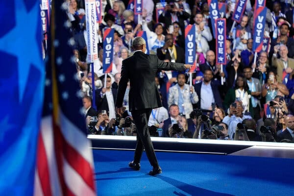 Former President Barack Obama walking on a blue-carpeted stage, his back to the camera, and pointing at the crowd. Former President Barack Obama walking on a blue-carpeted stage, his back to the camera, and pointing at the crowd.