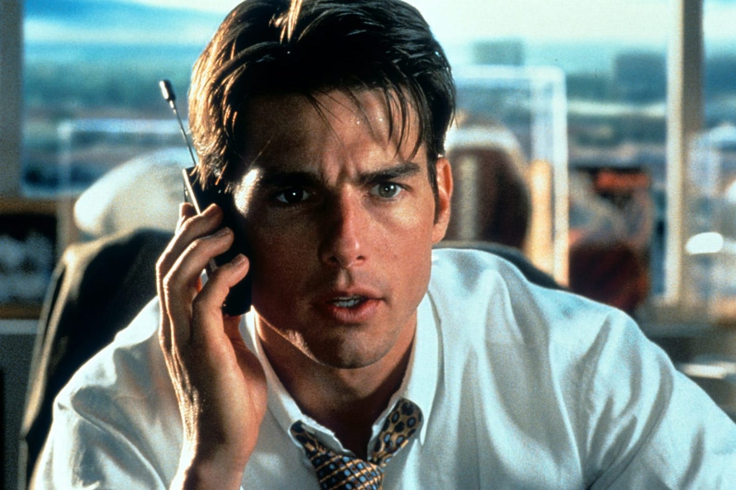 A picture from the movie Jerry Maguire. The titular character, a white guy in a white shirt with unbuttoned top collar and pulled down tie talks on a dated sell phone. The sweat on his forehead suggests that the guy is struggling. 
