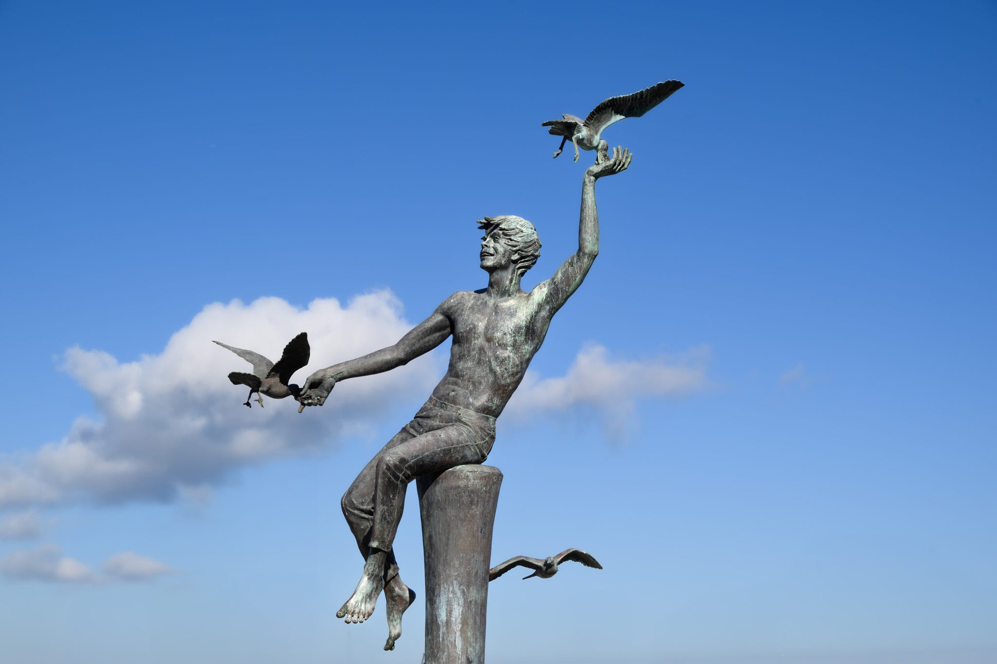 A sculpture of a young boy sitting on a pier piling feeding seagulls with one near each hand and another flying behind him; a blue sky and a white cloud in the background