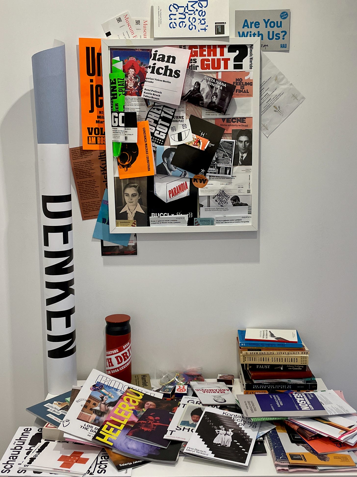 A cluttered desk with scattered books and magazines, and a corkboard filled with colourful flyers and papers on a white wall.