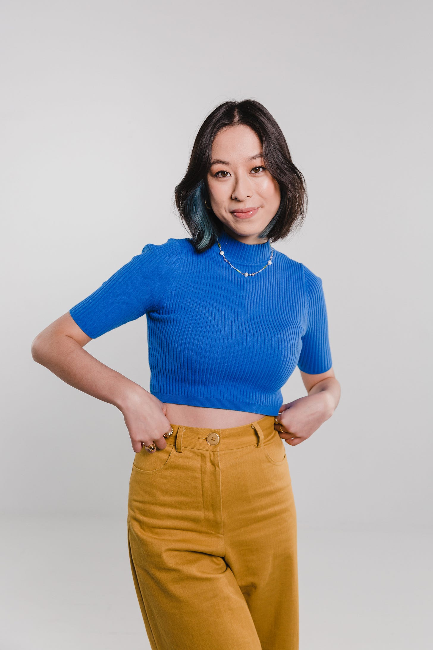 A young Asian Australian woman, standing in front of a gray backdrop and smiling, posing with her hands at her hips