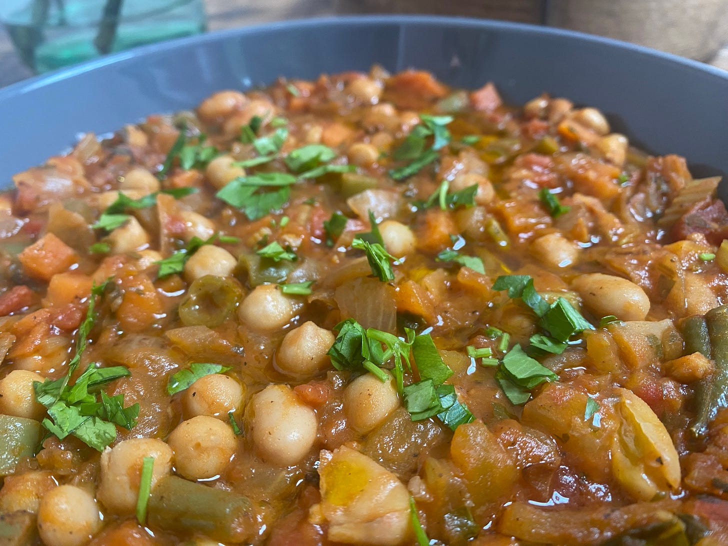 A grey bowl is on a table. In the bowl there are beans and vegetables in a tomato sauce and fresh herbs scattered on top
