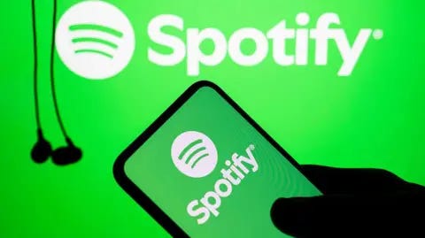 Getty Images The silhouette of a hand holds a phone with the green and white Spotify logo on it. In the background hang some black wired earbuds.