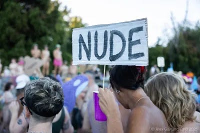 A person holds a sign reading “NUDE” above their head in a crowd of people at Denny Blaine Park, many appearing nude or topless, during a protest event. Trees and cardboard puppets are visible in the background. A person holds a sign reading “NUDE” above their head in a crowd of people at Denny Blaine Park, many appearing nude or topless, during a protest event. Trees and cardboard puppets are visible in the background.
