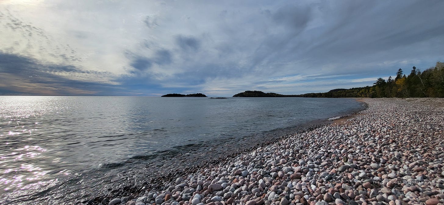 A moody, wide-angle shot of a grey cobble beach at Gargantua Harbor. The sky is filled with dark, dramatic clouds, reflecting the rugged and remote nature of the Lake Superior Provincial Park wilderness.
