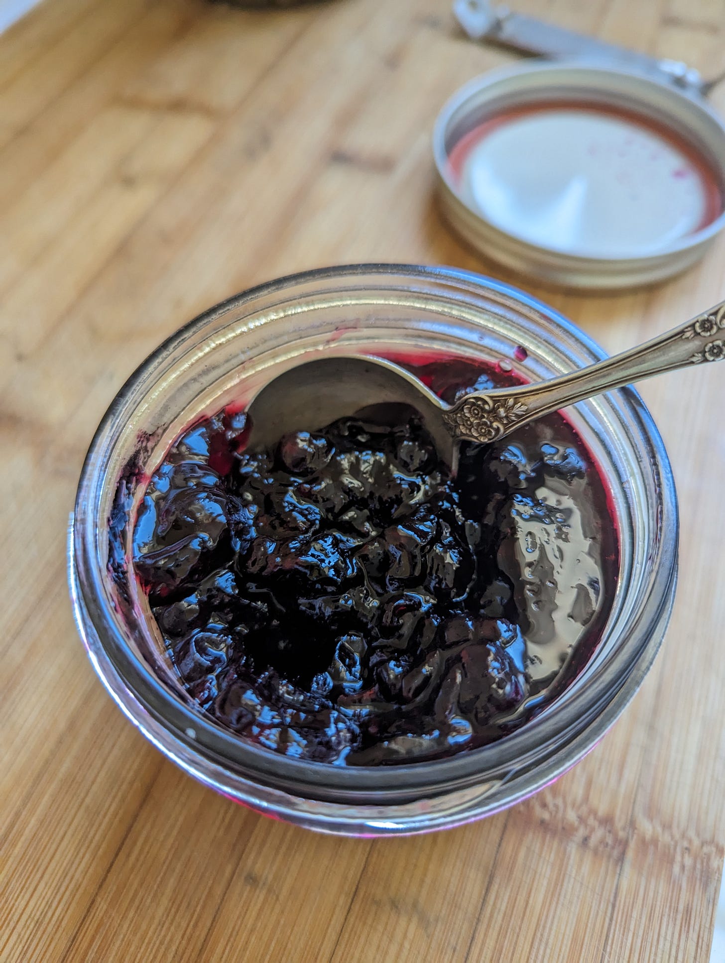 Half pint jar with black currant jam and a silver spoon. On a cutting board with a jar lid and ring in the background. 