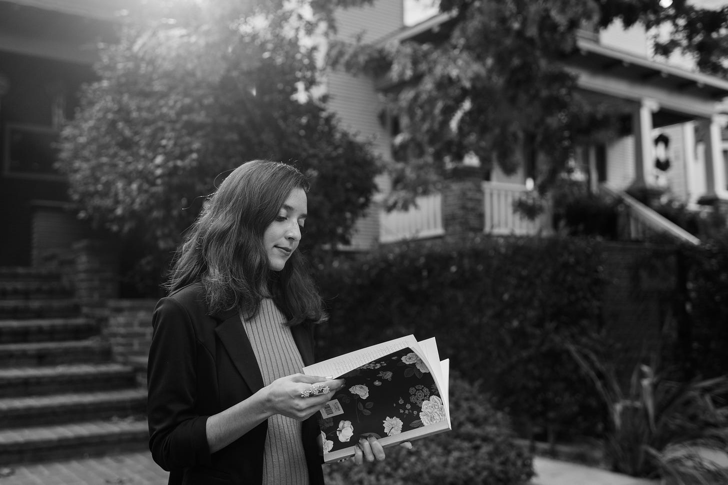 A black-and-white photo of a brown woman with short dark hair. She holds a floral black notebook and flips through its pages. The woman wears a black blazer and light dress. Behind her are trees and homes. A black-and-white photo of a brown woman with short dark hair. She holds a floral black notebook and flips through its pages. The woman wears a black blazer and light dress. Behind her are trees and homes.