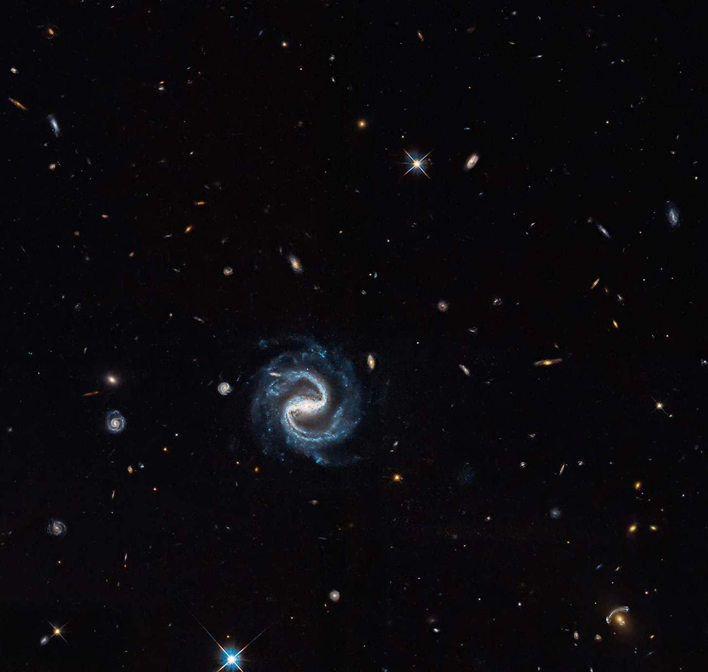 A spiral galaxy with two distinct arms forming a backwards "S" shape is in the foreground, against a background of many other much smaller, more distant galaxies. In the right lower corner, a gravitational lens can be seen, with a large, distinctive arc to the upper left.