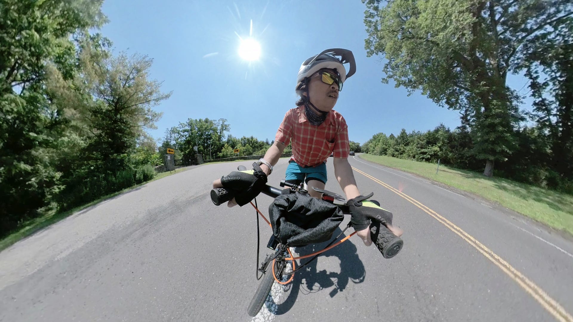 Photo of a man riding a bicycle. The camera is a couple of feet in front of the bicycle and point back towards the cyclist, who is leaning the bike left as he navigates a turn. There are trees on either side of the road and a blue sky with bright sun behind him.