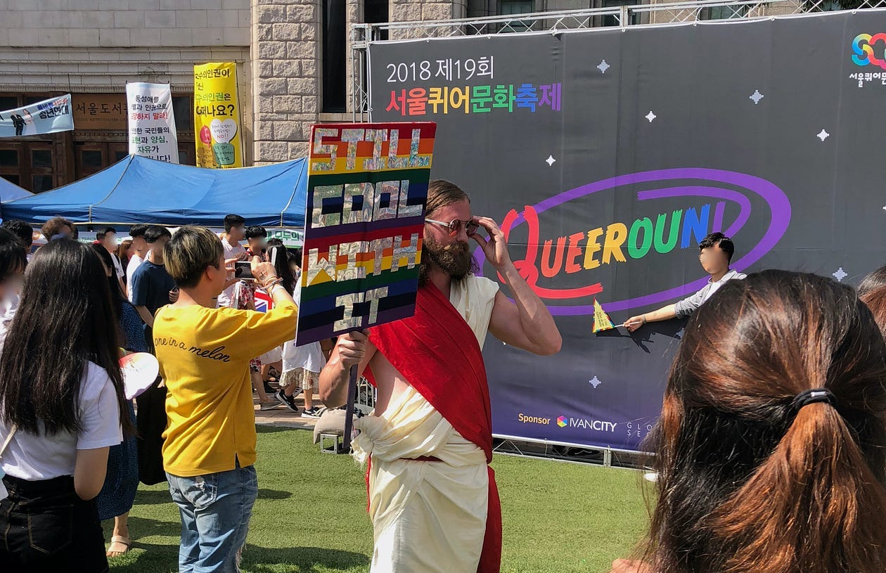 Jesus holds a rain sign saying “Still Cool With It” in front of Seoul Queer Festival goers
