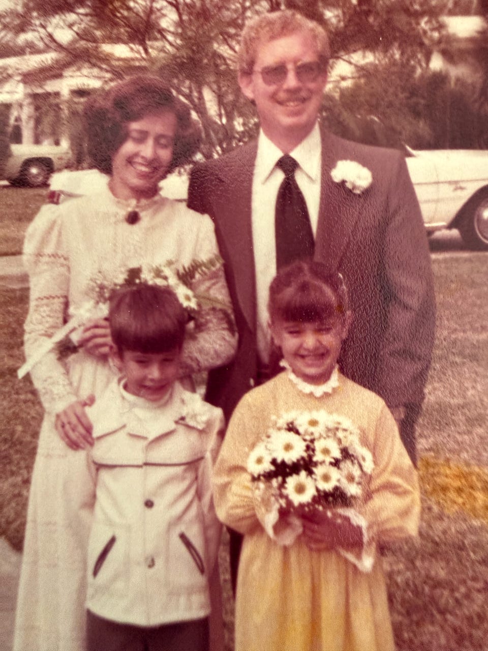 A man and woman, both dressed formally and holding flowers, stand outside with two young children, a boy and a girl, who are also dressed up and holding bouquets. A man and woman, both dressed formally and holding flowers, stand outside with two young children, a boy and a girl, who are also dressed up and holding bouquets.