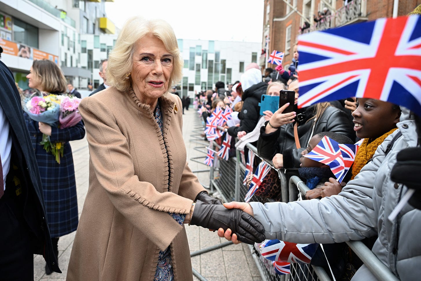 Queen Camilla shaking hands with people waiting outside a community centre wearing a biege long coat and leather gloves Queen Camilla shaking hands with people waiting outside a community centre wearing a biege long coat and leather gloves