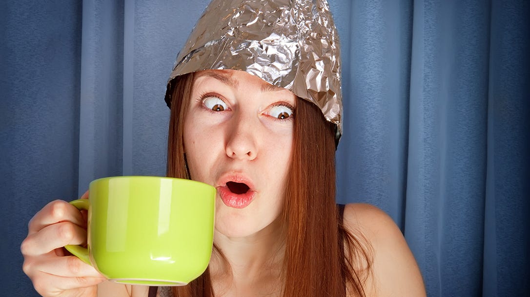 Young woman in tinfoil hat in front of computer holds bright green coffee mug and has comical stunned expression at what she's viewing.